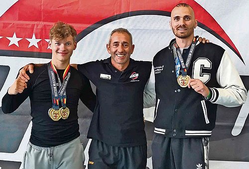Zu sehen sind drei Männer, die in die Kamera lächeln. In der Mitte steht der Trainer, außen rechts und links die beiden Sportler mit ihren Medaillen.