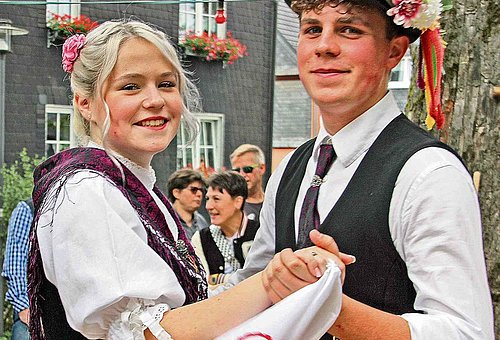 Das Foto zeigt ein Kirchweihpaar in Tracht. Beide schauen in die Kamera. Der Mann trägt einen Hut mit Blumenschmuck, das Mädchen hat ein weißes, besticktes Taschentuch in der rechten Hand. Sie stehen in Tanzstellung.