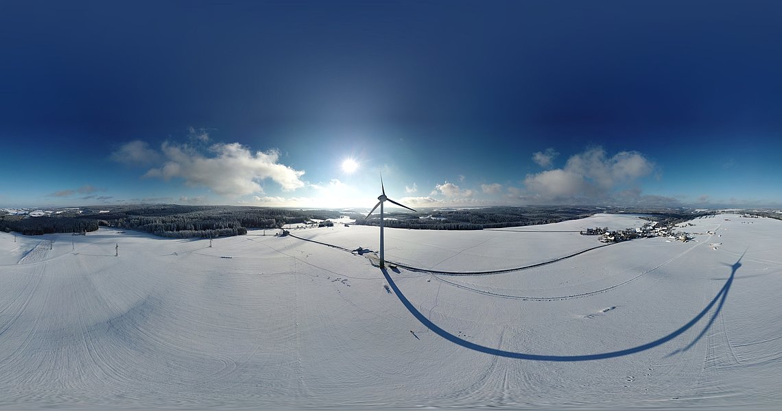 Das Foto zeigt eine Panoramansicht Lauenhains im Winter. Es liegt Schnee und der Himmel ist blau mit nur wenigen Wolken. Das Windrad ist deutlich in der Mitte zu sehen. 