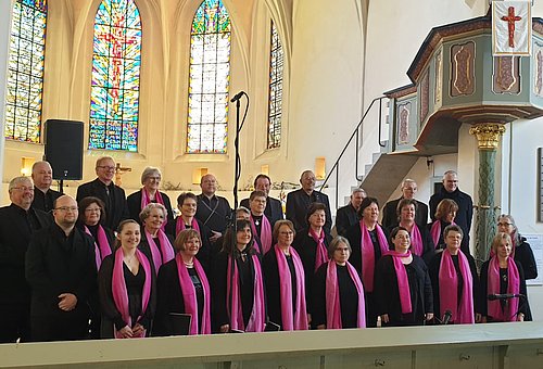 Ein gemischter Chor in einer Kirche. Alle tragen schwarze Kleidung, die Frauen zusätzlich ein pinkfarbenes Halstuch.