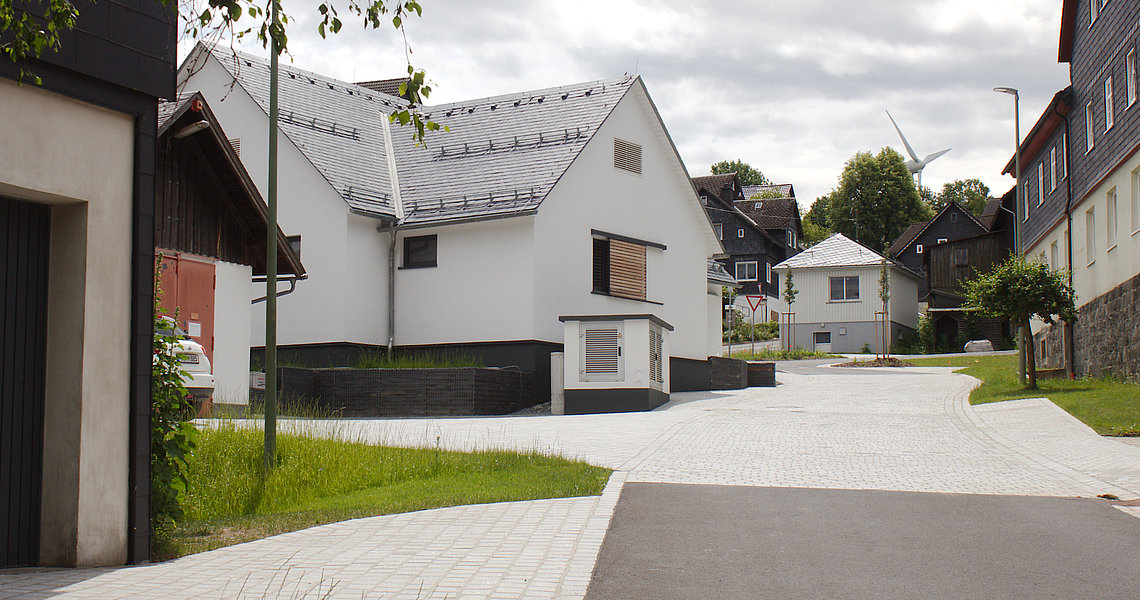 Zentral im Bild ist das neue Dorfgemeinschaftshaus Lauenhain zu sehen. Die Wände sind weiß verputzt, die Fensterläden sind aus Holzleisten und das Dach ist aus Schiefer. 