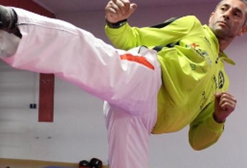 Im Bild ist ein Kampfsportler in Kampfposition zu sehen. Er trägt eine Trainingsanzug mit weißer Hose und grüner Jacke.