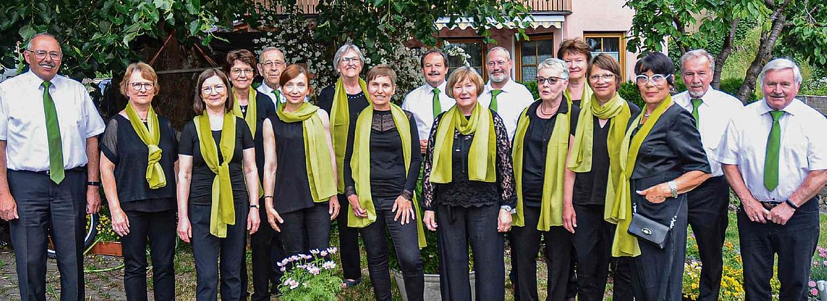 Das Bild zeigt den gesamten Chor. Die Frauen tragen schwarze Kleidung mit einem grünen Seidenschal, die Männer sind mit schwarzen Hosen, weißen Hemden und passenden grünen Krawatten gekleidet.