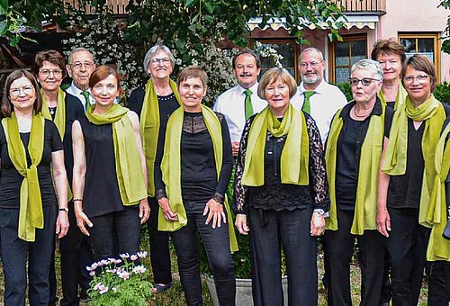 Das Bild zeigt den gesamten Chor. Die Frauen tragen schwarze Kleidung mit einem grünen Seidenschal, die Männer sind mit schwarzen Hosen, weißen Hemden und passenden grünen Krawatten gekleidet.