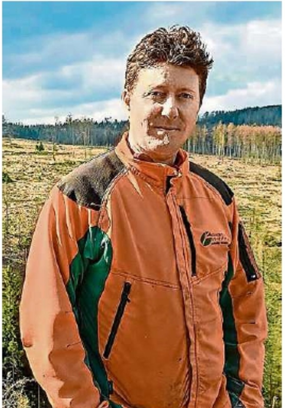 Ein Mann in einer orangefarbenen Waldjacke schaut in die Kamera. Er steht draußen und im Hintergrund sind abgeholzte Waldflächen.