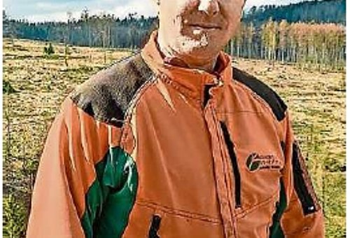 Ein Mann in einer orangefarbenen Waldjacke schaut in die Kamera. Er steht draußen und im Hintergrund sind abgeholzte Waldflächen. 