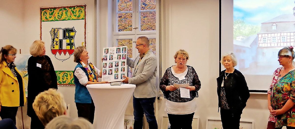 In einem Raum mit großen Gemälden an der Wand stehen 6 Frauen und ein Mann. Der Mann hält gemeinsam mit einer Frau eine Tafel mit vielen Porträitfotos von Menschen. Sie alle lächeln.