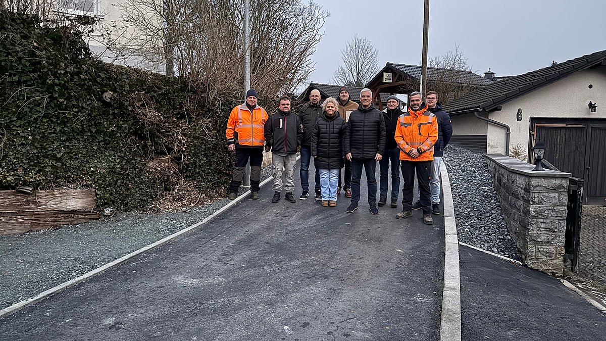 Eine Gruppe von Menschen stehen in einer Gruppe auf einer kleinen Straße. Im Hintergrund sind Wohnhäuser zu sehen. Alle lächeln in die Kamera.