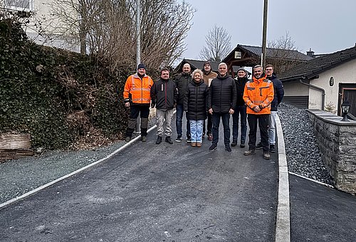 Eine Gruppe von Menschen stehen in einer Gruppe auf einer kleinen Straße. Im Hintergrund sind Wohnhäuser zu sehen. Alle lächeln in die Kamera.