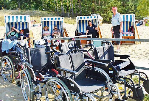 Im Vordergrund des Bildes sind leere Rollstühle zu sehen. Im Hintergrund sitzen alte Menschen entspannt in Strandkörben auf einem Sandstrand.