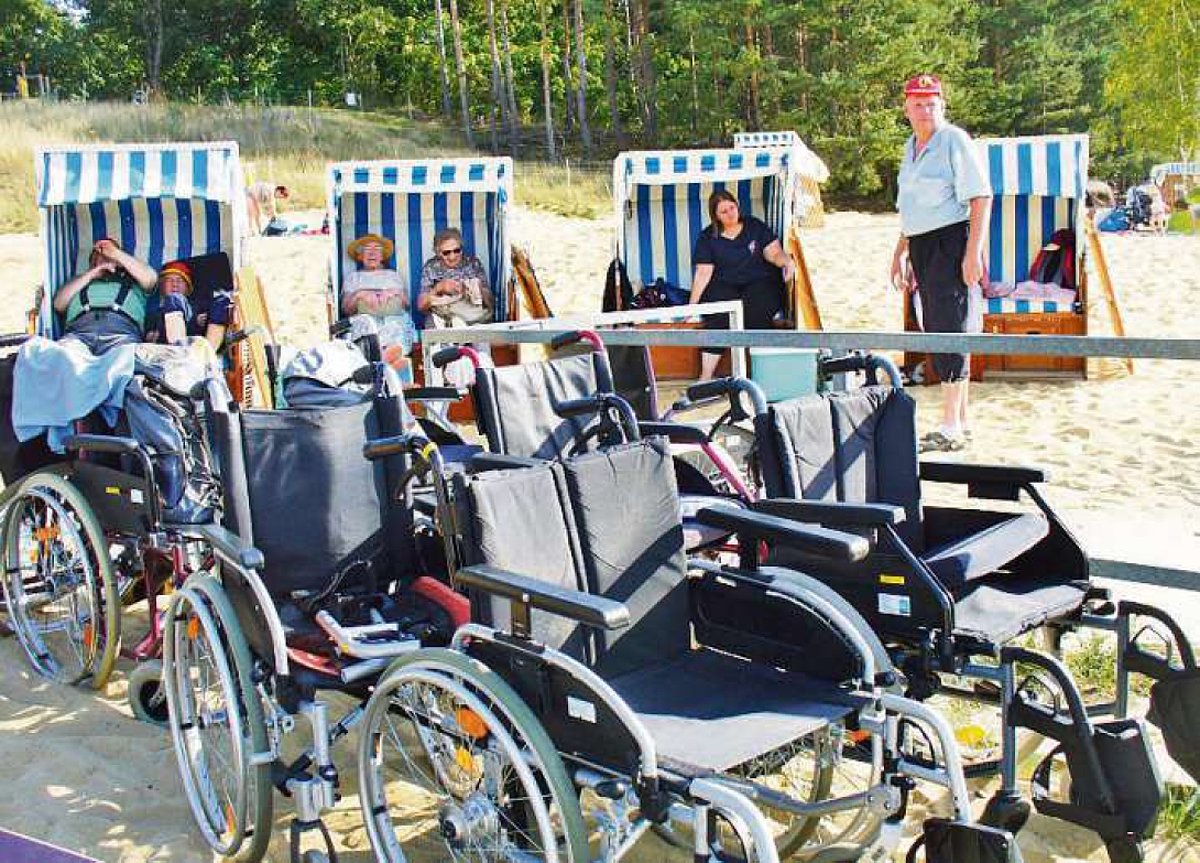 Im Vordergrund des Bildes sind leere Rollstühle zu sehen. Im Hintergrund sitzen alte Menschen entspannt in Strandkörben auf einem Sandstrand.