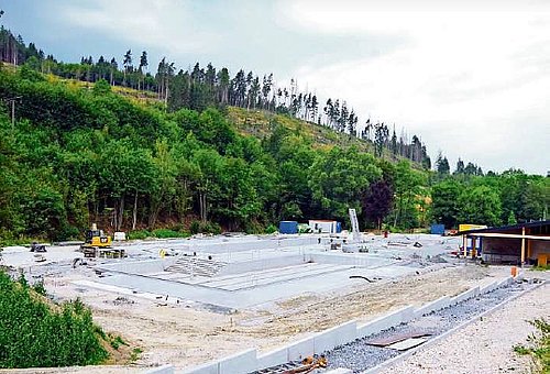 Ansicht einer Baustelle eines Freibades. Im Hintergrund sind bewaldete Hügel zu sehen. 
