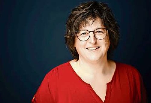 Eine Frau in Nahaufnahme. Sie steht vor einem dunklen Hintergrund. Die Frau hat eine rote Bluse an. Sie ist dunkelhaarig und trägt eine Brille. Sie lächelt in die Kamera.