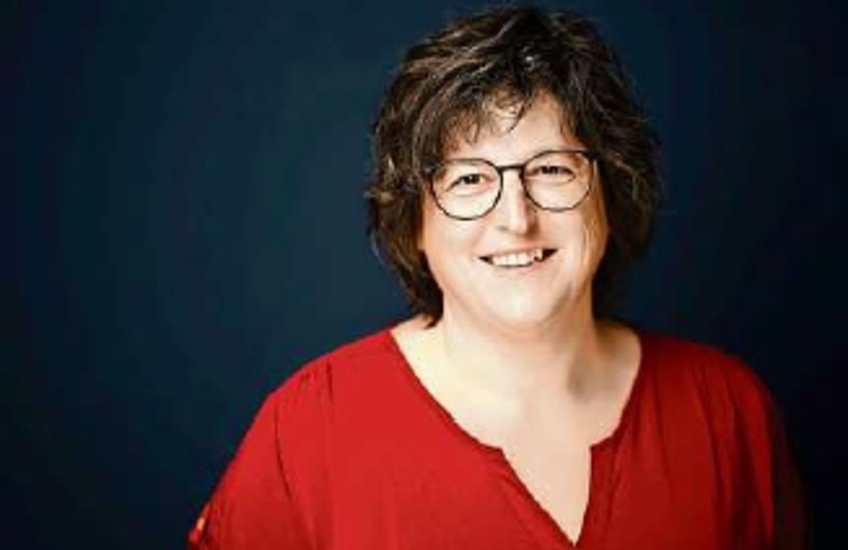 Eine Frau in Nahaufnahme. Sie steht vor einem dunklen Hintergrund. Die Frau hat eine rote Bluse an. Sie ist dunkelhaarig und trägt eine Brille. Sie lächelt in die Kamera.
