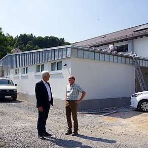 Zwei Männer stehen auf einer Baustelle. Im Hintergrund ist eine Flachbauhalle zu sehen. Es stehen Fahrzeuge herum.