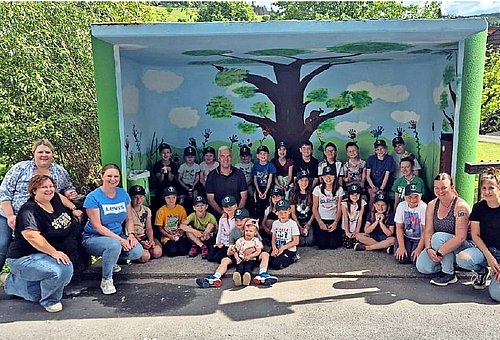 Vor einem gemalten Buswartehäuschen hocken viele Kinder und einige Erwachsene. Alle tragen Alltagskleidung und lächeln in die Kamera.