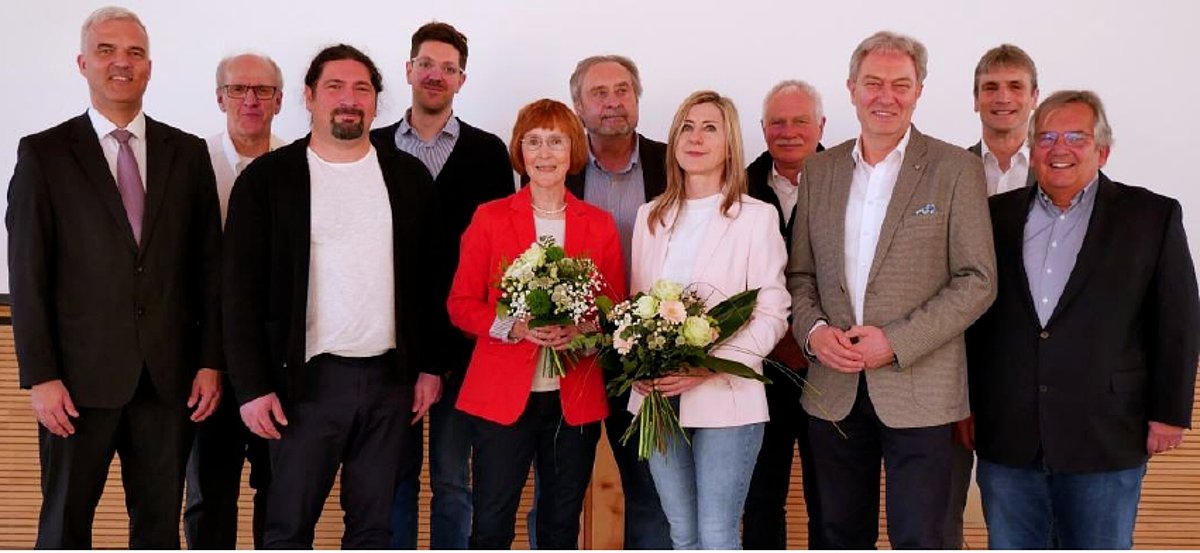 Eine Gruppe von Menschen, darunter zwei Frauen, stehen in zwei Reihen hintereinander und schauen in die Kamera.
