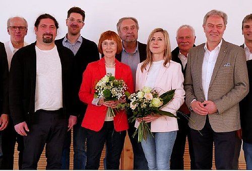 Eine Gruppe von Menschen, darunter zwei Frauen, stehen in zwei Reihen hintereinander und schauen in die Kamera.