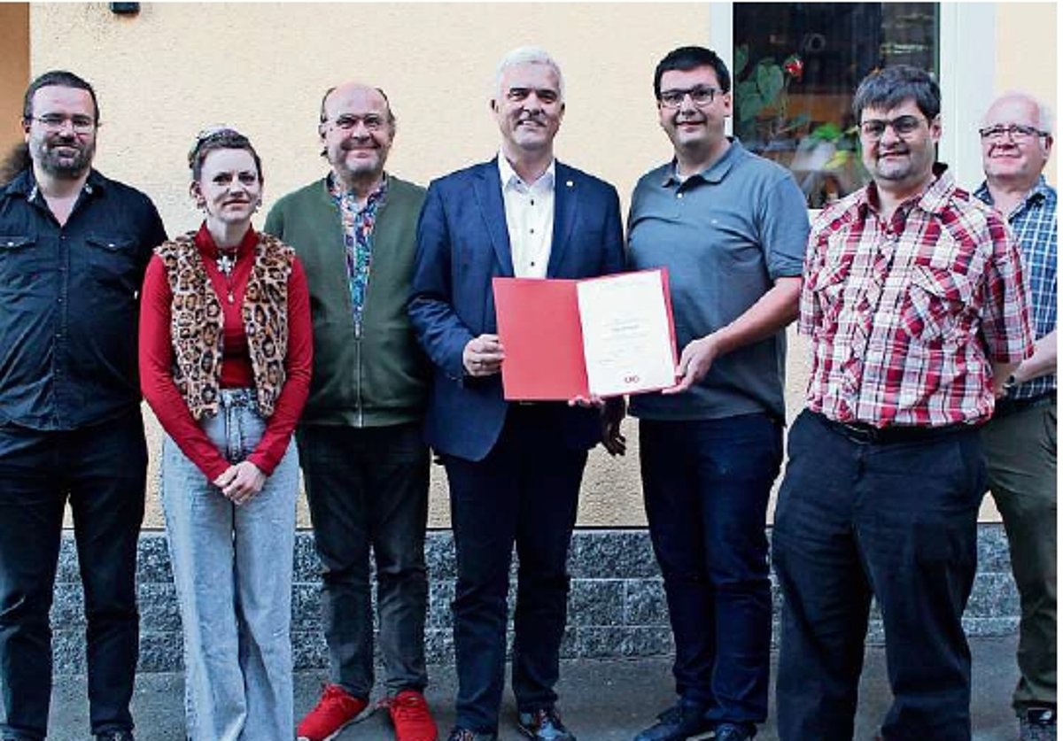 Eine Gruppe von Menschen, darunter eine Frau. In der Mitte halten zwei Männer eine rote Mappe mit einer Urkunde. Alle lächeln in die Kamera.