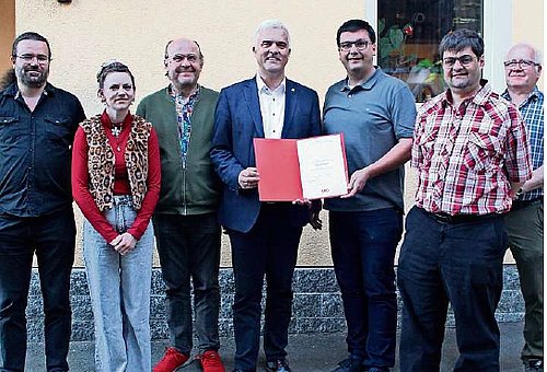 Eine Gruppe von Menschen, darunter eine Frau. In der Mitte halten zwei Männer eine rote Mappe mit einer Urkunde. Alle lächeln in die Kamera.