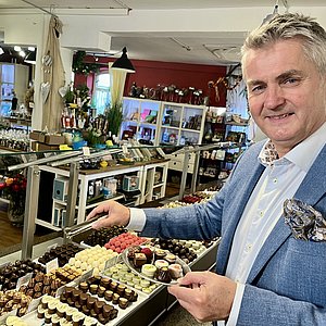 Ein Mann in Anzug steht in einem großen Verkaufsraum. Im Hintergrund sind helle Regale mit Pralinen und Dekorationen zu sehen. Der Mann lächelt in die Kamera. In einer Hand hält er eine kleine silberne Platte auf der verschiedenen Pralinen liegen. In der anderen Hand hält er eine silberne Zange. 