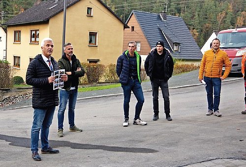 Mehrere Menschen stehen in einem Halbkreis auf einer Straße. Sie hören dem Redner zu, das ist Bürgermeister Timo Ehrhardt. Im Hintergrund sind Wohnhäuser zu sehen. 