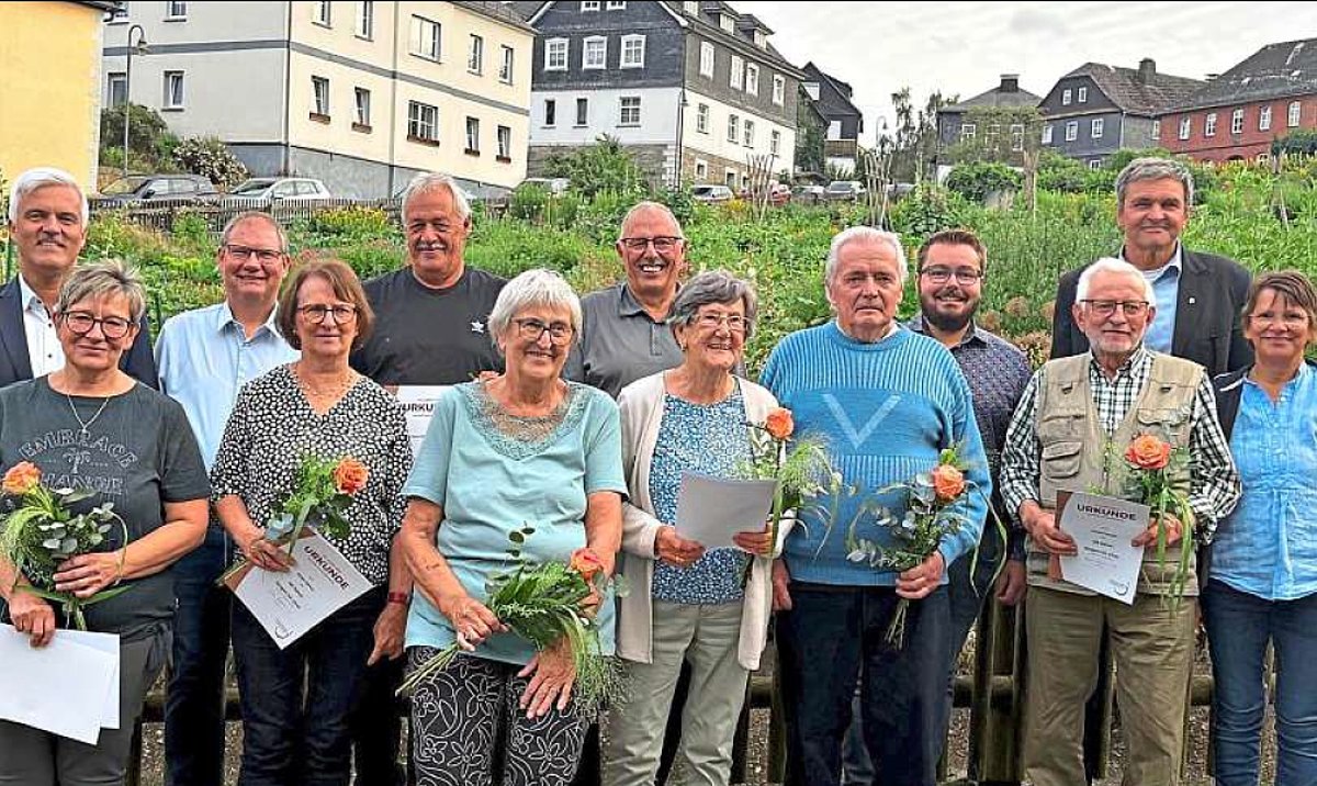 Eine Gruppe von Menschen verschiedenen Alters stehen in einer Gruppe zusammen. Die Personen in der vorderen Reihe halten je einen kleinen Blumenstrauß und eine Urkunde in ihren Händen. Alle lächeln in die Kamera. Im Hintergrund ist ein großer Garten mit umliegenden Häusern zu sehen.