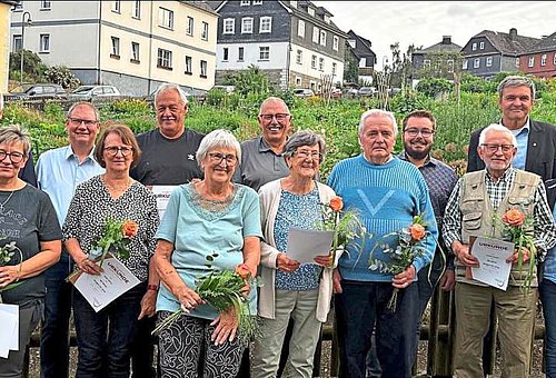 Eine Gruppe von Menschen verschiedenen Alters stehen in einer Gruppe zusammen. Die Personen in der vorderen Reihe halten je einen kleinen Blumenstrauß und eine Urkunde in ihren Händen. Alle lächeln in die Kamera. Im Hintergrund ist ein großer Garten mit umliegenden Häusern zu sehen.