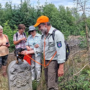 Das Foto zeigt eine Wandergruppe mit 3 Frauen, einem Mann und einem Dackel. Alle schauen zu den Hund, der auf einem hohen Grenzstein sitzt. 