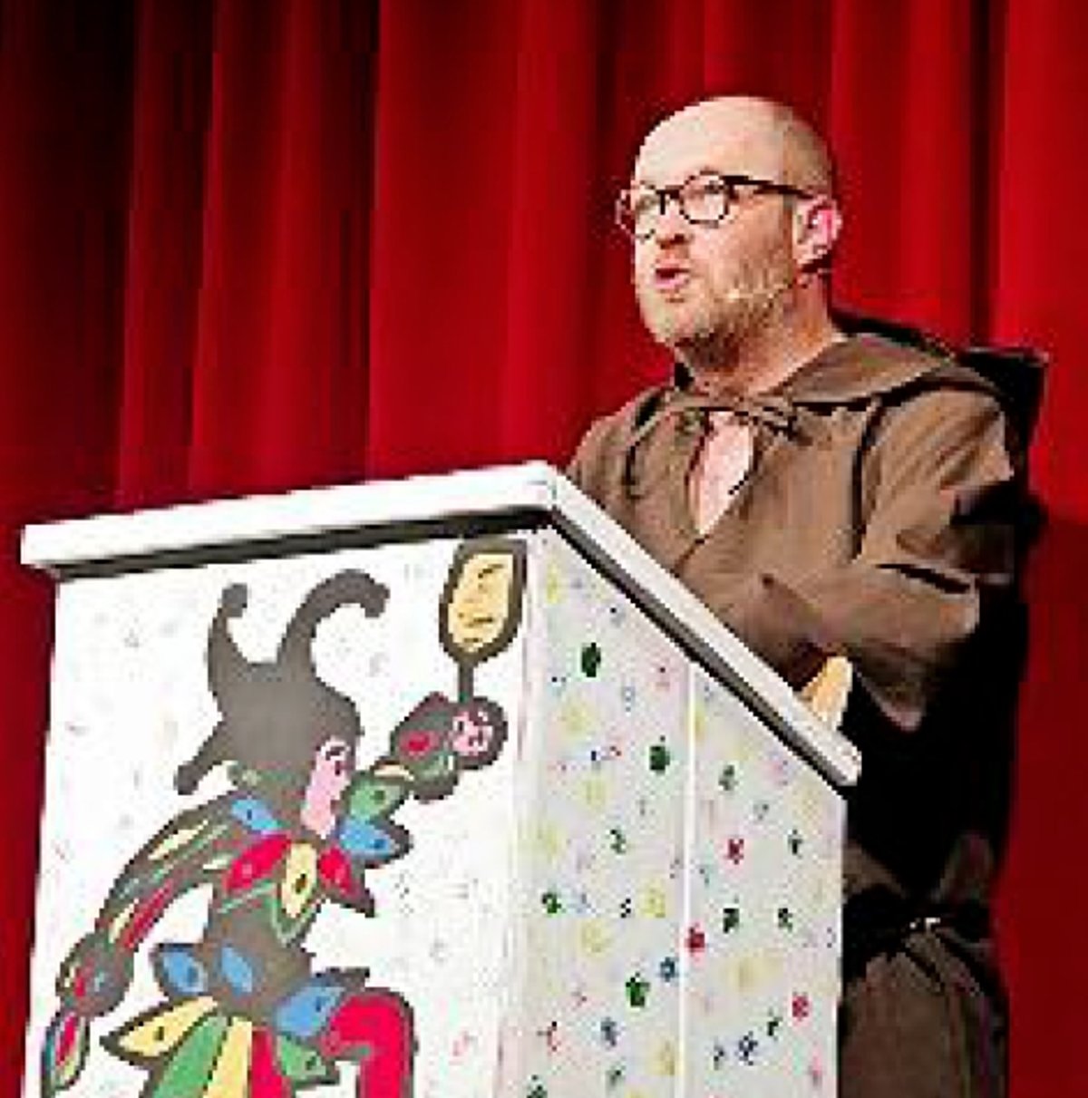 Ein Mann in einem Mönchskostüm steht an einem Rednerpult, auf dem ein Til Eulenspiegel gemalt ist.