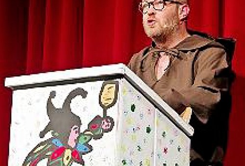 Ein Mann in einem Mönchskostüm steht an einem Rednerpult, auf dem ein Til Eulenspiegel gemalt ist.
