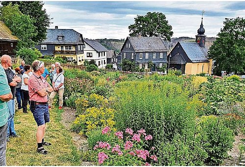 Ein blühender Garten. Im Hintergrund Wohnhäuser und eine kleine Kirche. Am Bildrand stehen mehrere Menschen und schauen in die Ferne.