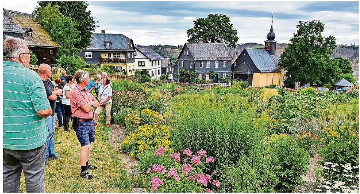 Ein blühender Garten. Im Hintergrund Wohnhäuser und eine kleine Kirche. Am Bildrand stehen mehrere Menschen und schauen in die Ferne.