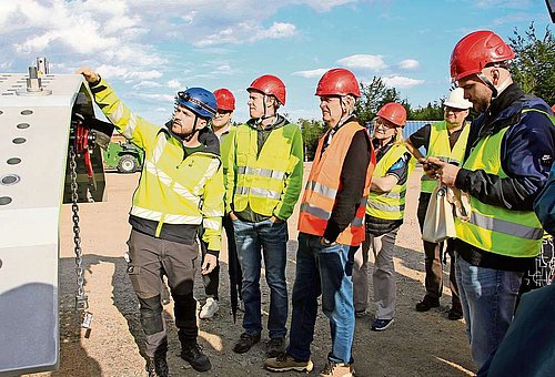 Mit roten Helmen und gelben Warnwesten bestaunt eine Gruppe von Männern und einer Frau die Baustelle. Links auf dem Bild ist ein großes Stahlteil für den Bau der Windräder zu sehen.