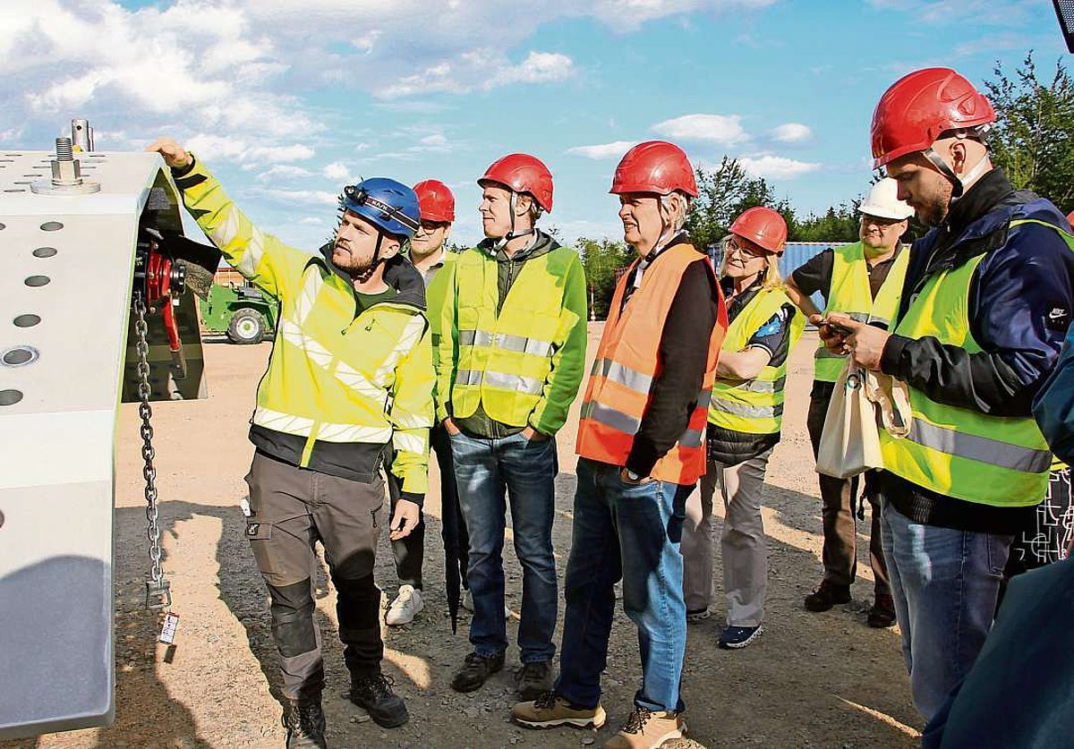 Mit roten Helmen und gelben Warnwesten bestaunt eine Gruppe von Männern und einer Frau die Baustelle. Links auf dem Bild ist ein großes Stahlteil für den Bau der Windräder zu sehen.