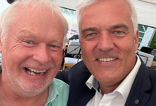 Selfie von zwei Männern. Beide haben graue Haare und lachen in die Kamera.