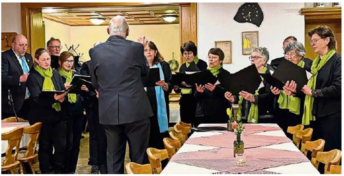 In einem Raum stehen Frauen und Männer in schwarzer Kleidung zusammen. Sie tragen grüne Tücher bzw. blaue Krawatten und halten alle eine Mappe in der Hand. Der Dirigent in schwarzem Anzug steht vor ihnen und ist nur von hinten zu sehen.