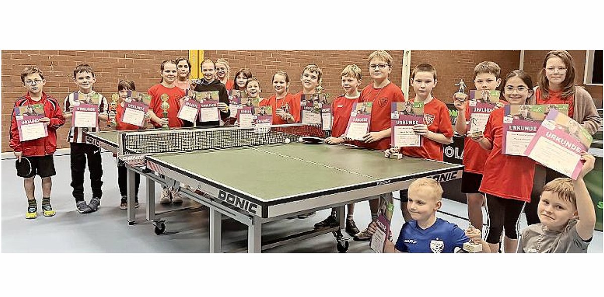 In einer Turnhalle stehen um eine Tischtennisplatte sehr viele Kinder in Sportkleidung. Sie stehen, knieen oder hocken. Alle halten Urkunden in ihrer Hand und lachen in die Kamera.