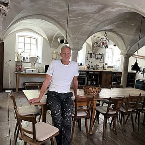 In einem Gewölbekeller steht ein Mann in Arbeitskleidung. Er lehnt an einem großen Esstisch, der den Raum dominiert. Der Raum ist eine Küche mit Schränken und Küchenmobiliar.