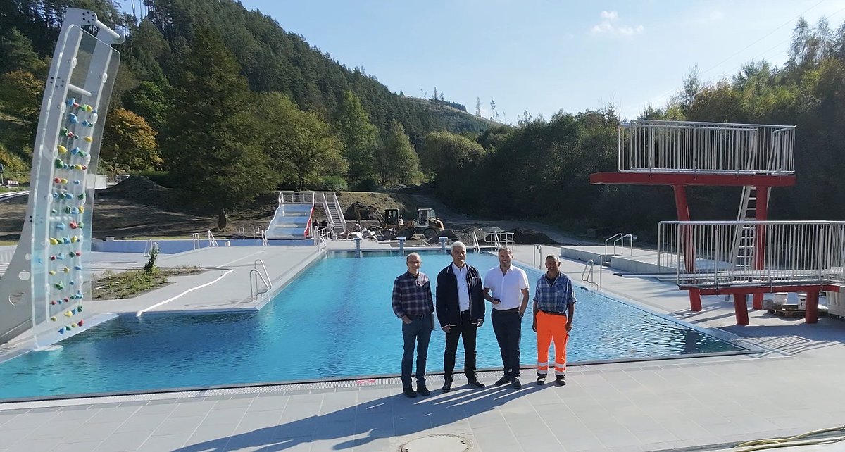 Vor einem gefüllten Freibad-Schwimmerbecken stehen vier Männer. Sie stehen mit dem Rücken zum Becken und schauen in die Kamera.