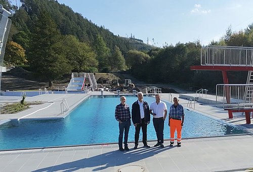 Vor einem gefüllten Freibad-Schwimmerbecken stehen vier Männer. Sie stehen mit dem Rücken zum Becken und schauen in die Kamera.