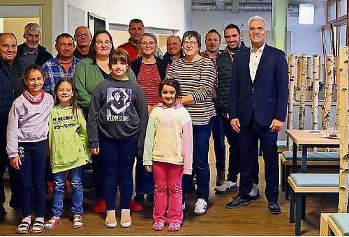 In einem Raum steht eine große Gruppe von Menschen, darunter auch vier Kinder. Sie sind alle verschieden gekleidet und schauen in die Kamera. Am rechten Bildrand sieht man neue Tische und Sitzbänke, dazwischen stehen Birkenstämme als Raumteiler.