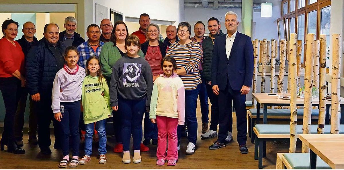 In einem Raum steht eine große Gruppe von Menschen, darunter auch vier Kinder. Sie sind alle verschieden gekleidet und schauen in die Kamera. Am rechten Bildrand sieht man neue Tische und Sitzbänke, dazwischen stehen Birkenstämme als Raumteiler.