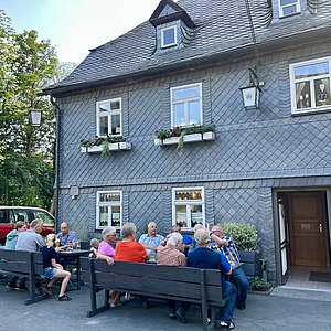 Ein komplett verschiefertes Haus mit Obergeschoss. Das Haus hat weiße Fensterrahmen und eine dunkle Holztür. Vor dem Haus stehen Bänke und Tisch für die Gäste. Menschen sitzen sich zugewandt an den Tischen.