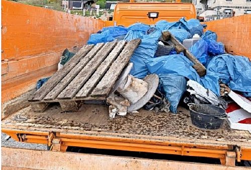 Blick auf die Ladefläche einen orangefarbenen LKW. Darauf sind zahlreiche blaue gefüllte Müllsäcke und viel Unrat zu sehen. 