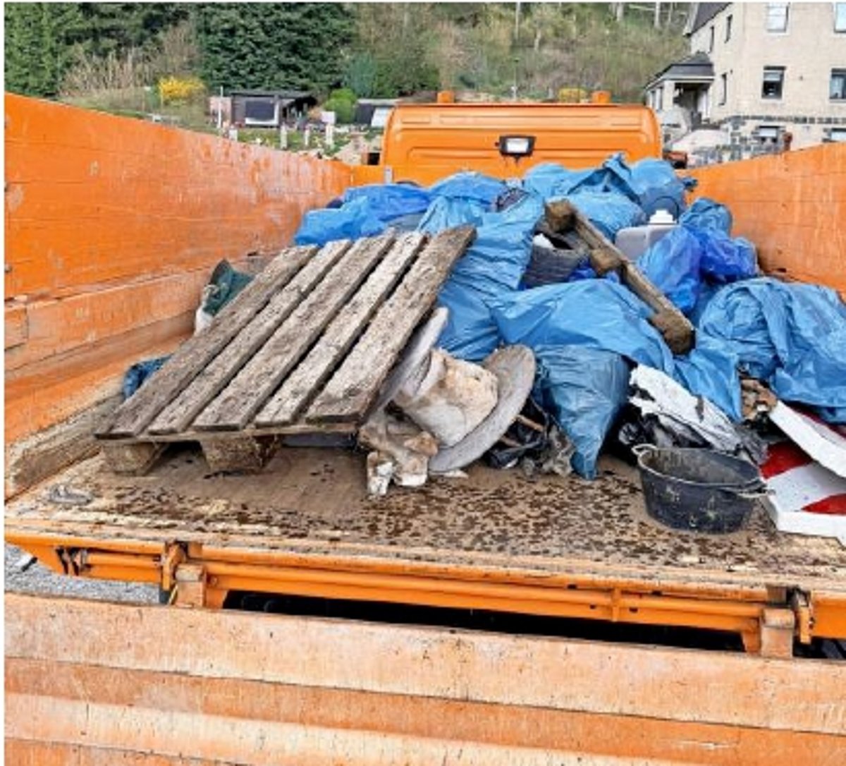 Blick auf die Ladefläche einen orangefarbenen LKW. Darauf sind zahlreiche blaue gefüllte Müllsäcke und viel Unrat zu sehen.