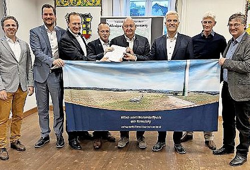 Acht Männer stehen nebeneinander und halten ein Banner vor sich. Darauf ist ein Windrad zu sehen. Die Männer, alle in Anzügen, lächeln in die Kamera.