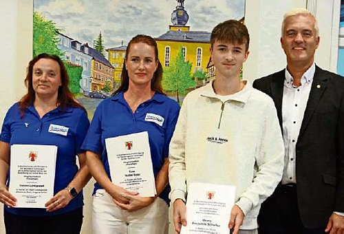 zwei Frauen mit blauen Poloshirts, ein junger Mann im weißen Langarmshirt und Bürgermeister Timo Ehrhardt im Anzug stehen in einem Gebäude vor einer Wand mit einem Gemälde mit Kirche. Die zwei Frauen und der junge Mann halten je eine Urkunde in der Hand. Alle lächeln in die Kamera.