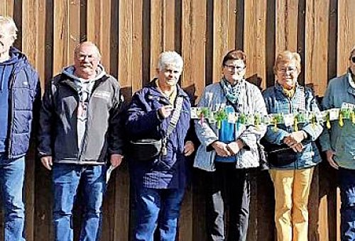 Drei Männer und drei Frauen stehen nebeneinander an einer Holzwand und halten Geldscheine in den Händen.