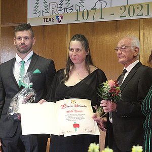 Vier Menschen stehen nebeneinander auf der Bühne. Gezeigt wird die Urkunde, die Willi Rimpl bekommt. Er steht zwischen den beiden Frauen.